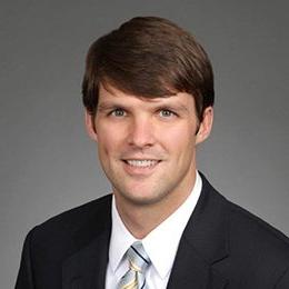 Person standing in front of grey background wearing dark suit