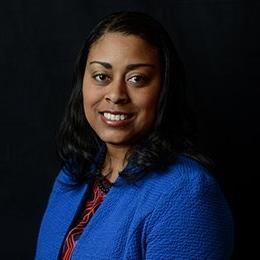 Person wearing royal blue jacket standing in front of dark background smiling