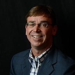 Person wearing navy suit coat in front of dark background smiling 