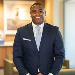 Man wearing navy suit and light blue tie standing inside library